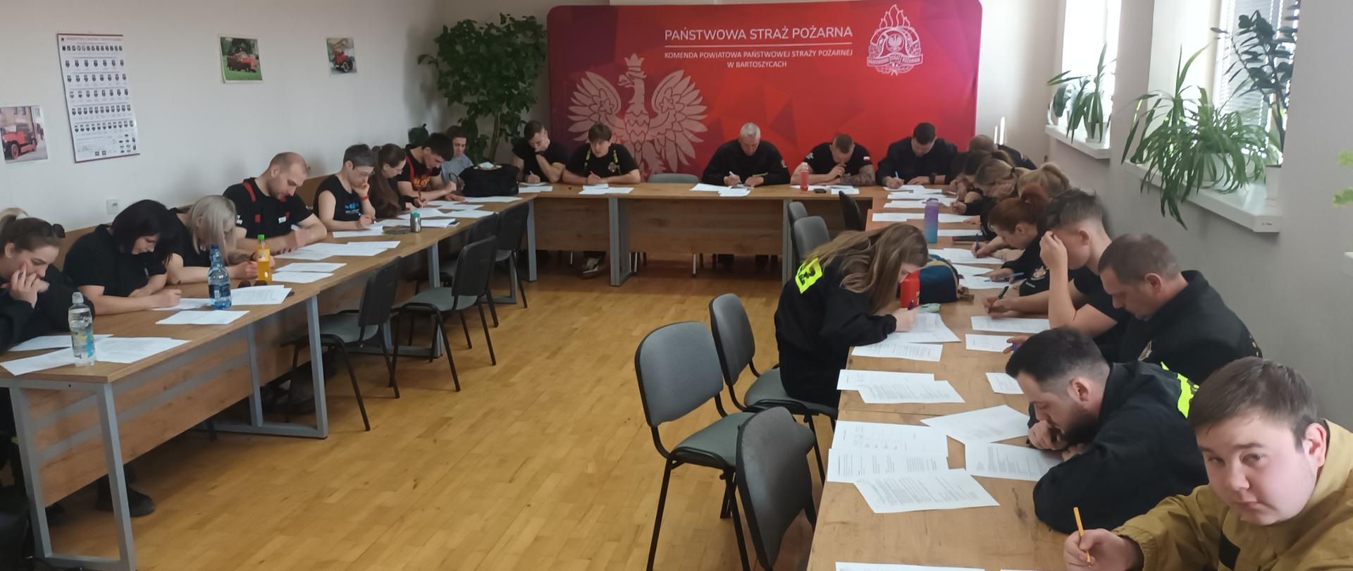Zdjęcie przedstawia salę szkoleniową, w której kilkudziesięciu uczestników w ubraniach oznaczonych jako „STRAŻ” i „OSP” pisze test pisemny. Pomieszczenie należy do Komendy Powiatowej Państwowej Straży Pożarnej w Bartoszycach, co potwierdza widoczna na ścianie plansza z godłem i nazwą instytucji. Uczestnicy siedzą przy ustawionych w podkowę stołach, koncentrując się na wypełnianiu arkuszy egzaminacyjnych. Wnętrze jest jasne, z drewnianą podłogą, dużymi oknami po jednej stronie i dekoracją roślinną. Zdjęcie dokumentuje etap szkolenia lub egzaminu teoretycznego dla strażaków.