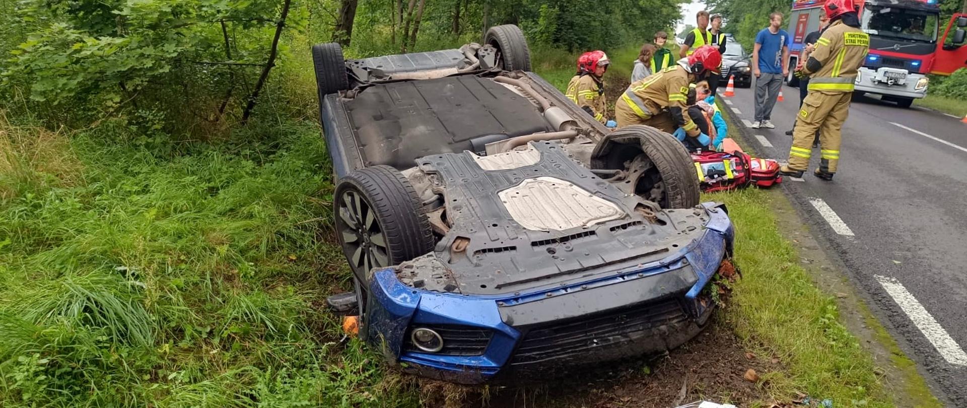 Ten sam samochód przewrócony na dach leży na poboczu, a obok strażacy pomagają osobie poszkodowanej, leżącej na noszach. W tle widać wóz strażacki i kilku ratowników medycznych. Droga jest częściowo zablokowana pachołkami.