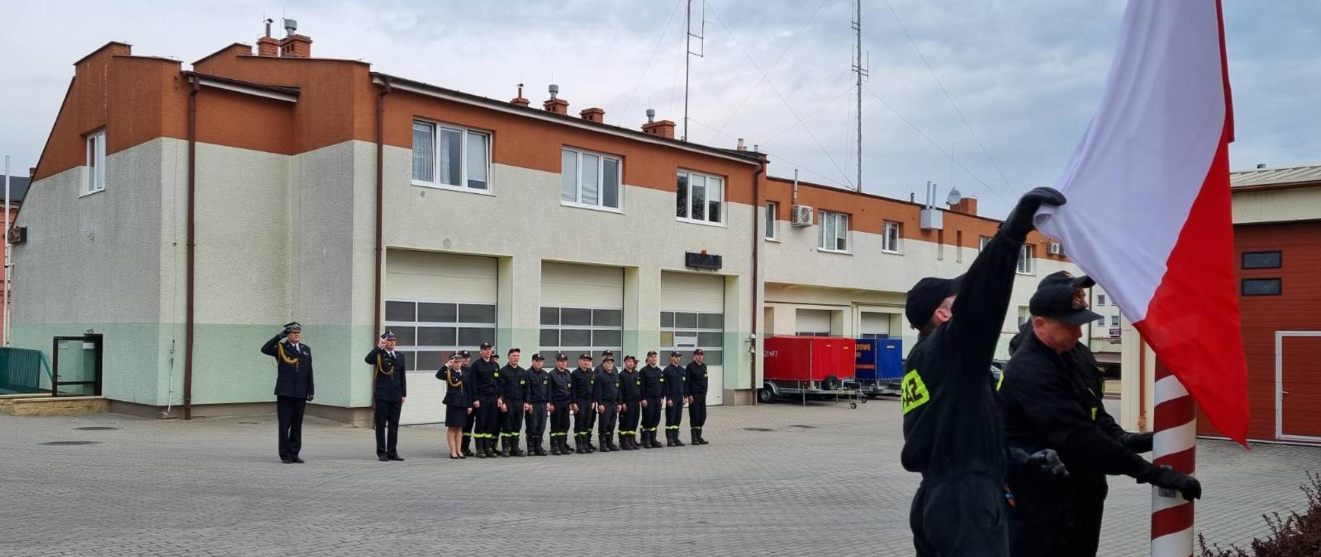 Zdjęcie przedstawia moment podniesienia flagi państwowej na maszt, w tle strażacy i kierownictwo Komendy oddają hołd