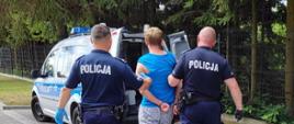 Zdjęcie przedstawia funkcjonariuszy policji prowadzących zatrzymanego do policyjnego samochodu