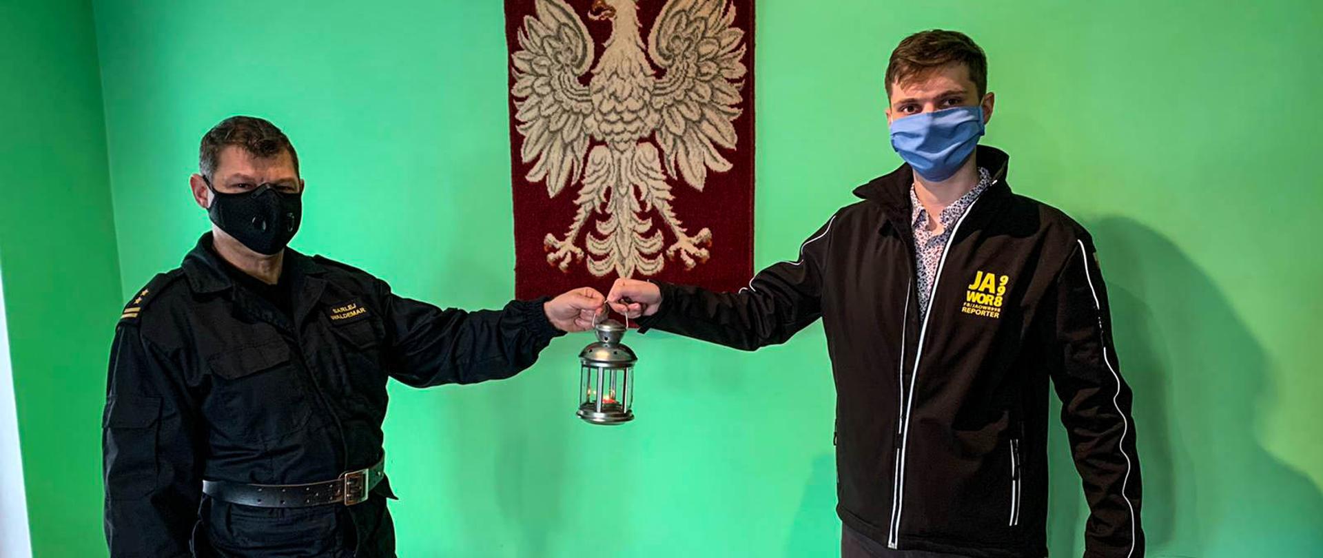 Obraz przedstawia Komendanta Powiatowego Państwowej Straży Pożarnej w Jaworze brygadiera Waldemara Sarleja otrzymującego lampion z Betlejemskim Światłem Pokoju od redaktora portalu Jawor998 Piotra Tłuściaka. W tle zielona ściana na której wisi godło państwowe.