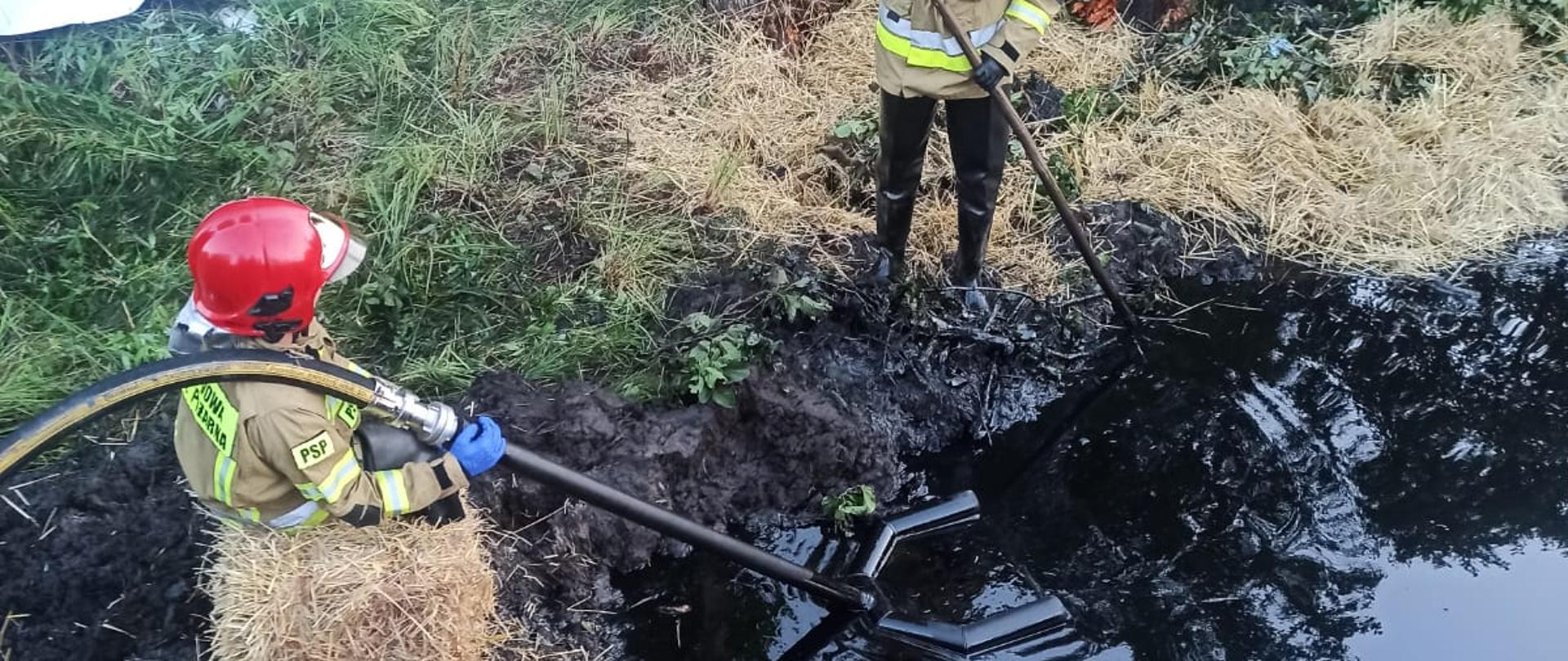 Zdjęcie obrazuje strażaków zbierających ciecz ropopochodną z powierzchni zbiornika wodnego. Używają do tego celu skimera. Jedne z nich z powodu bardzo grząskiego gruntu musiał usiąść na kostce słomy aby się nie zapaść do gruntu. W tle widać roślinność i złamane drzewo.