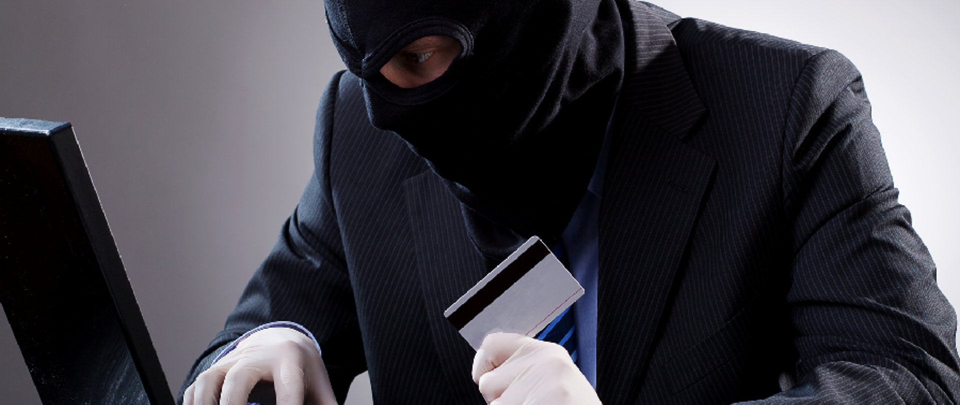 Zamaskowana osoba przy komputerze, w dłoni karta płatnicza