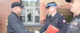 Zdjęcie przedstawia strażaków podczas uroczystej zmiany służby.
W tle budynki.
