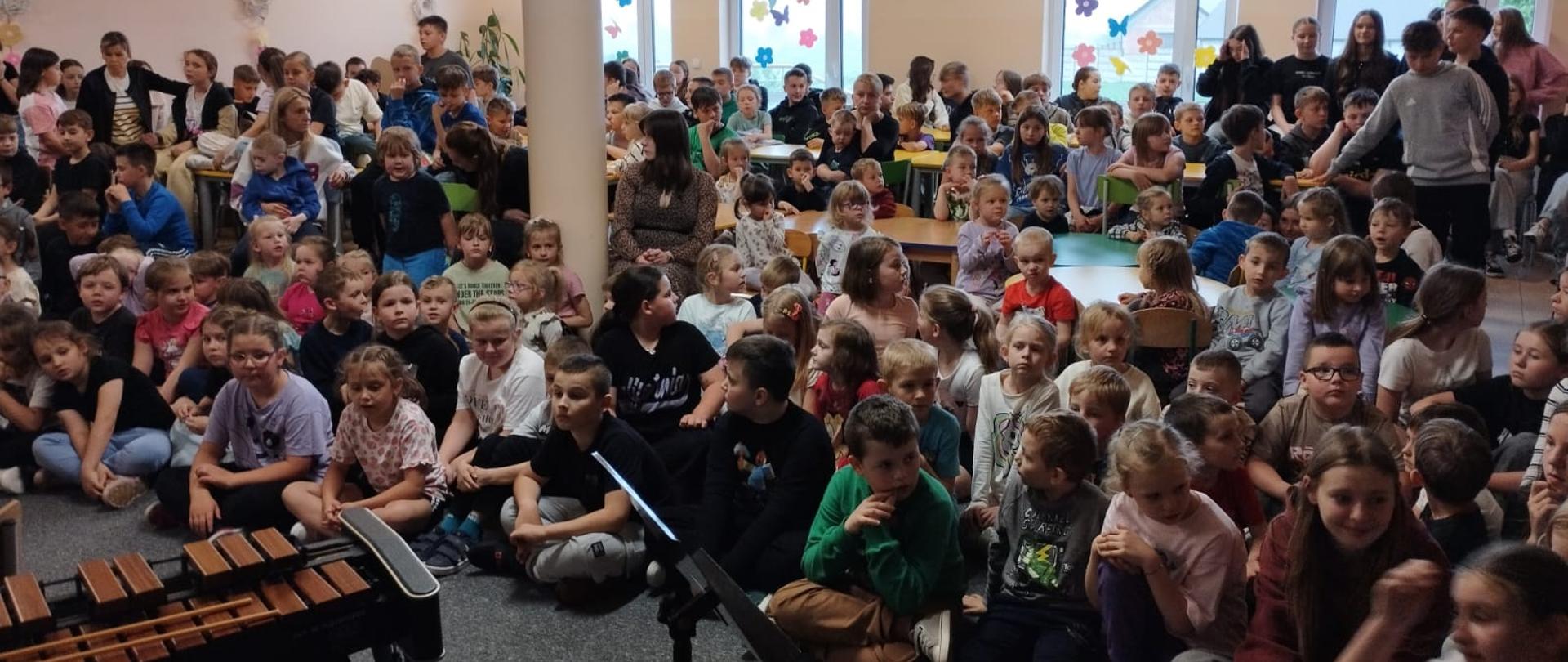 Na zdjęciu widać dużą grupę dzieci siedzących w sali. Są zgromadzone na podłodze, a w tle znajdują się okna z widokiem na zewnątrz. W sali panuje radosna atmosfera, a dzieci są ubrane w różne kolorowe ubrania. Na pierwszym planie znajduje się instrument muzyczny, prawdopodobnie wibrafon. Wygląda na to, że jest to wydarzenie związane z muzyką lub występ.