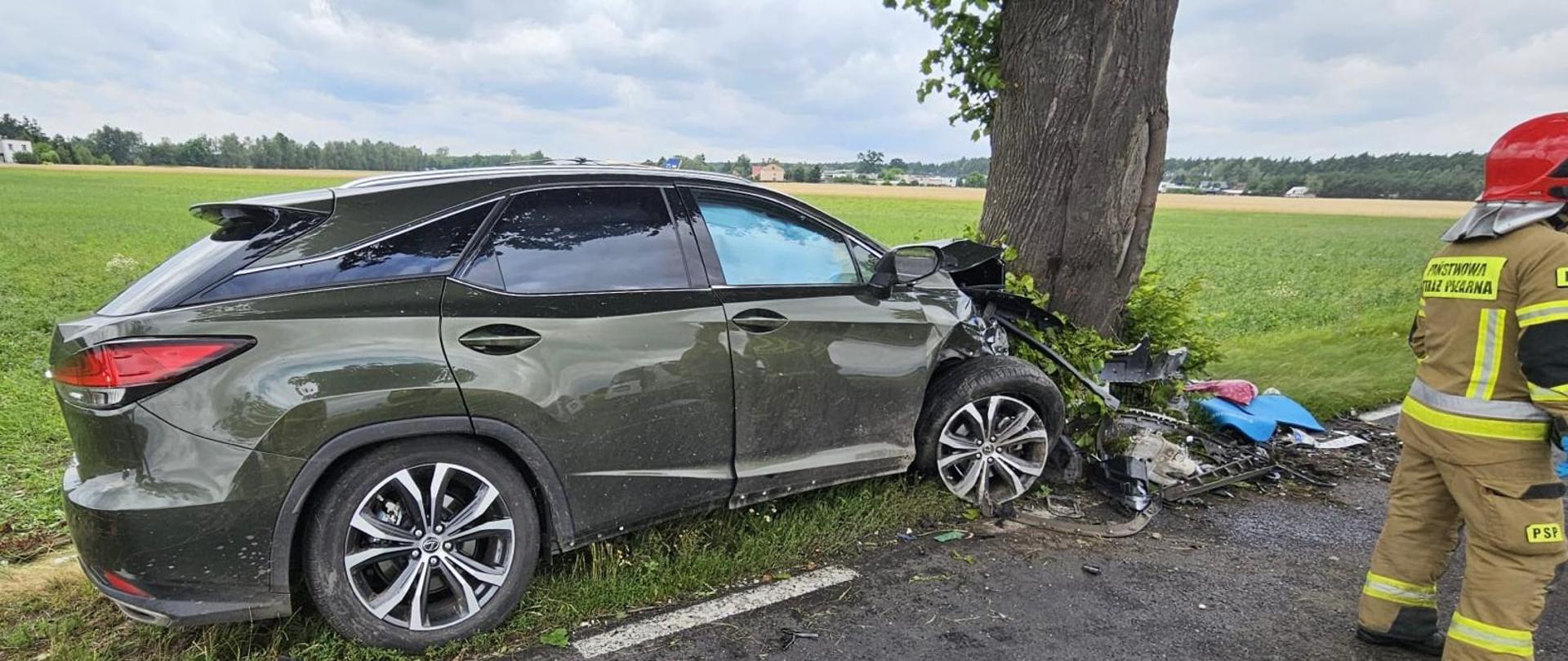 Samochód SUV z rozbitym przodem stoi przy drzewie po wypadku drogowym. Obok znajdują się strażacy zabezpieczający miejsce zdarzenia