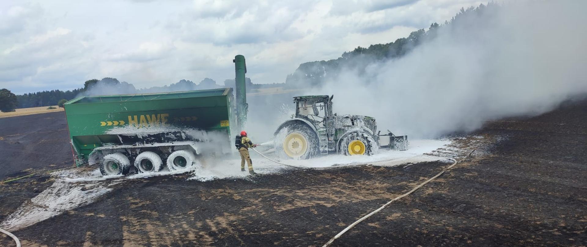Zdjęcie przedstawia moment gaszenia ciągnika rolniczego z przyczepą, które gasi strażak podający piane gaśniczą.