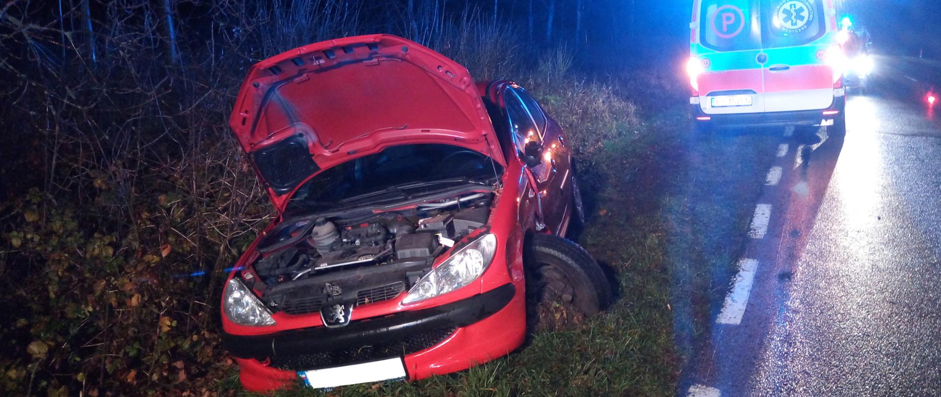Samochód marki Peugeot stojący w przydrożnym rowie po zderzeniu z innym samochodem osobowym. W pojeździe uszkodzony bok od strony kierowcy. W tle ambulans pogotowia ratunkowego.