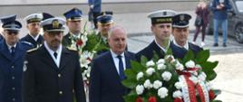 Wojewoda Pomorski oraz funkcjonariusze służ mundurowych idą w trzyosobowych rzędach obok siebie przed nimi idzie żołnierz Marynarki Wojennej który niesie wieniec.