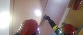 Na zdjęciu widać dwóch ratowników PSP w ubraniach specjalnych, aparatach oddechowych oraz hełmach z kamerą termowizyjną przed wejściem do pomieszczenia w którym jest pożar.