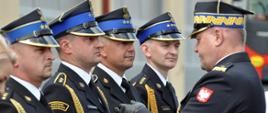 Zdjęcie zrobione na zewnątrz podczas uroczystości. Funkcjonariusz Państwowej Straży Pożarnej w stopniu generała wręcza odznaczenie strażakowi, który jest ustawiony w szeregu razem z innymi strażakami odbierającymi odznaczenia. 
