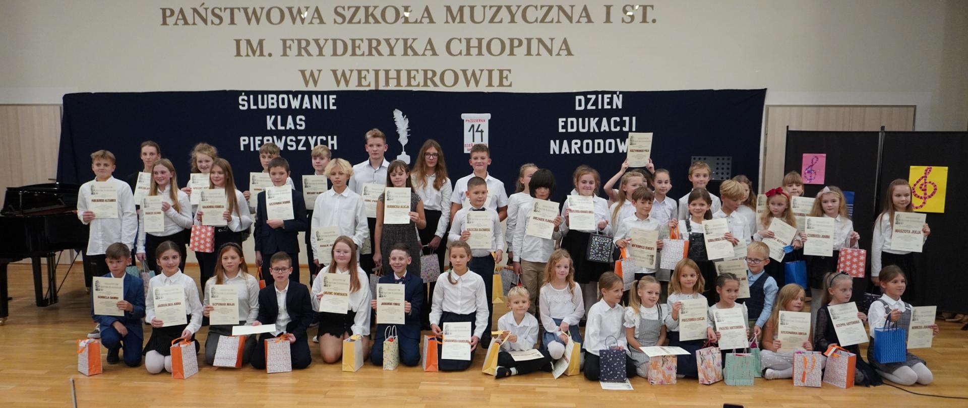 Na scenie pozują do zdjęcia uczniowie klas pierwszych. Trzymają w dłoni pamiątkowe dyplomy oraz torebki z upominkami.