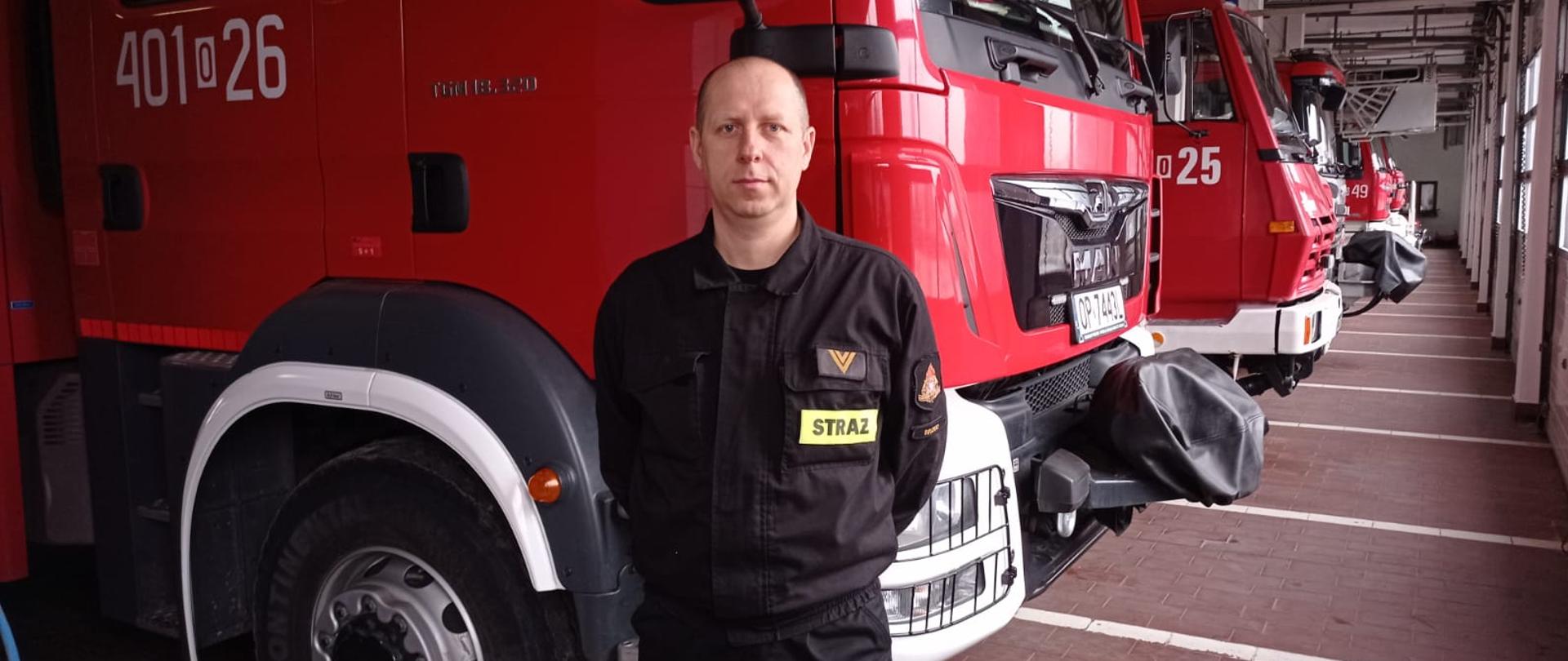 Zdjęcie przedstawia st.ogn. Marcina Garnczarczyka - strażaka JRG Brzeg stojącego na tle samochodów pożarniczych na terenie garażu jednostki.