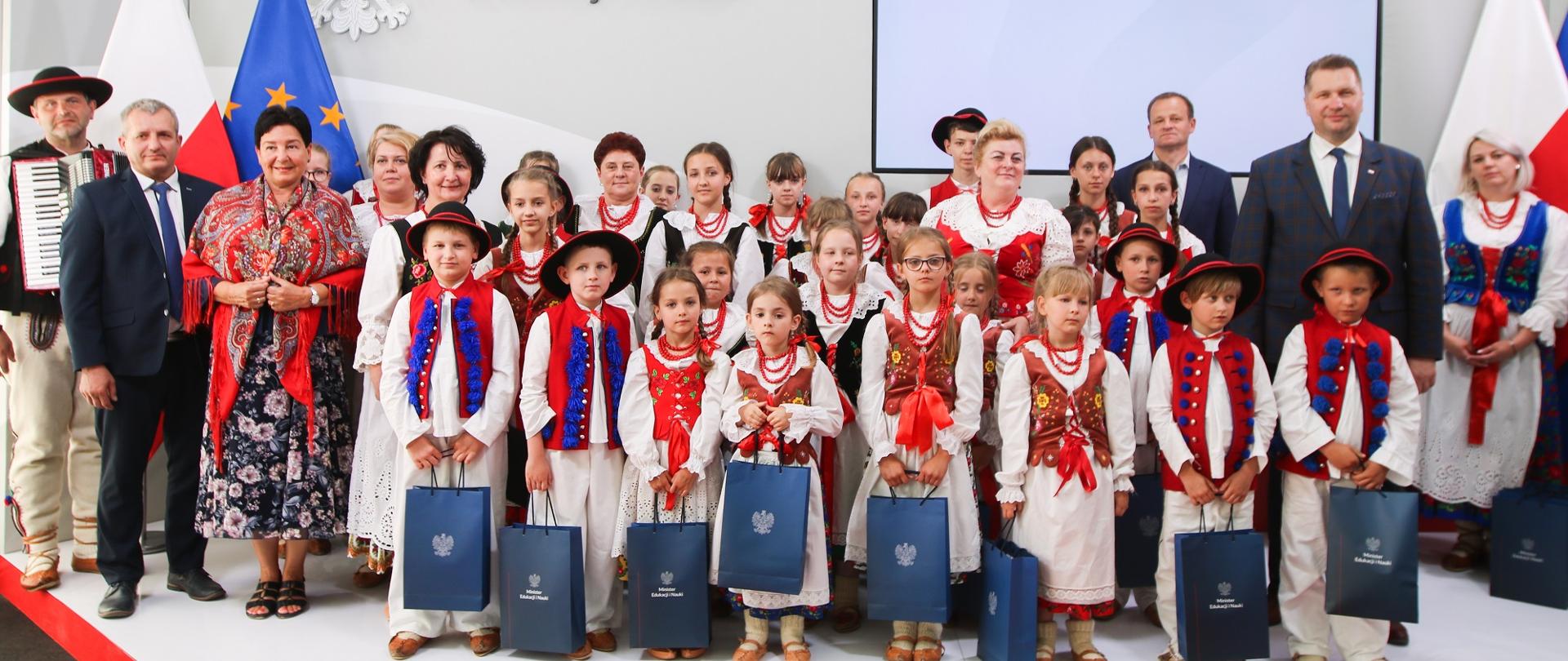 Grupa dzieci w ludowych strojach stoi na podwyższeniu, trzymają w rękach papierowe torby, za nimi stoi minister Czarnek i kilka innych osób.