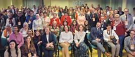 Zdjęcie grupowe. Uczestnicy konferencji pozują do wspólnego zdjęcia w sali konferencyjnej. Część uczestników siedzi w dwóch rzędach na krzesłach, pozostali stoją za nimi. W tle widać żółte ściany sali konferencyjnej.