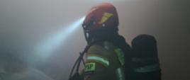 Na pierwszym planie fotografii znajduje się strażak wyposażony w sprzęt ochrony układu oddechowego oraz linię wężową, który podaje prąd wody na palące się materiały. W tle widać silne zadymienie uniemożliwiające rozpoznanie pomieszczenia oraz strumień światła