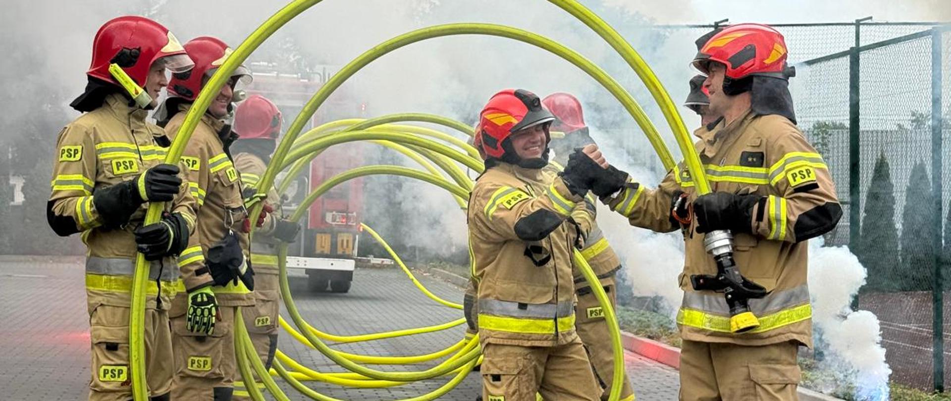 Strażacy tworzą szpaler, trzymając w rękach zielone węże pożarnicze uniesione w kształt łuków. Jeden ze strażaków przechodzi środkiem, uśmiechnięty, witając się ze stojącymi kolegami. W tle widoczny jest dym, nadający uroczysty charakter wydarzeniu.