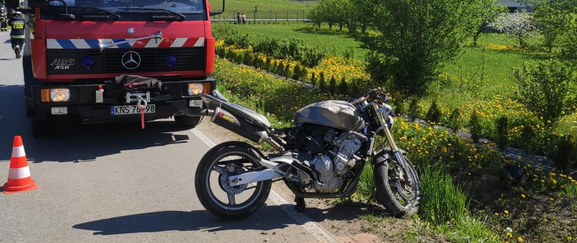 Motocykl na drodze na kołach przed nim elementy motocyklisty. Za motocyklem samochód pożarniczy.