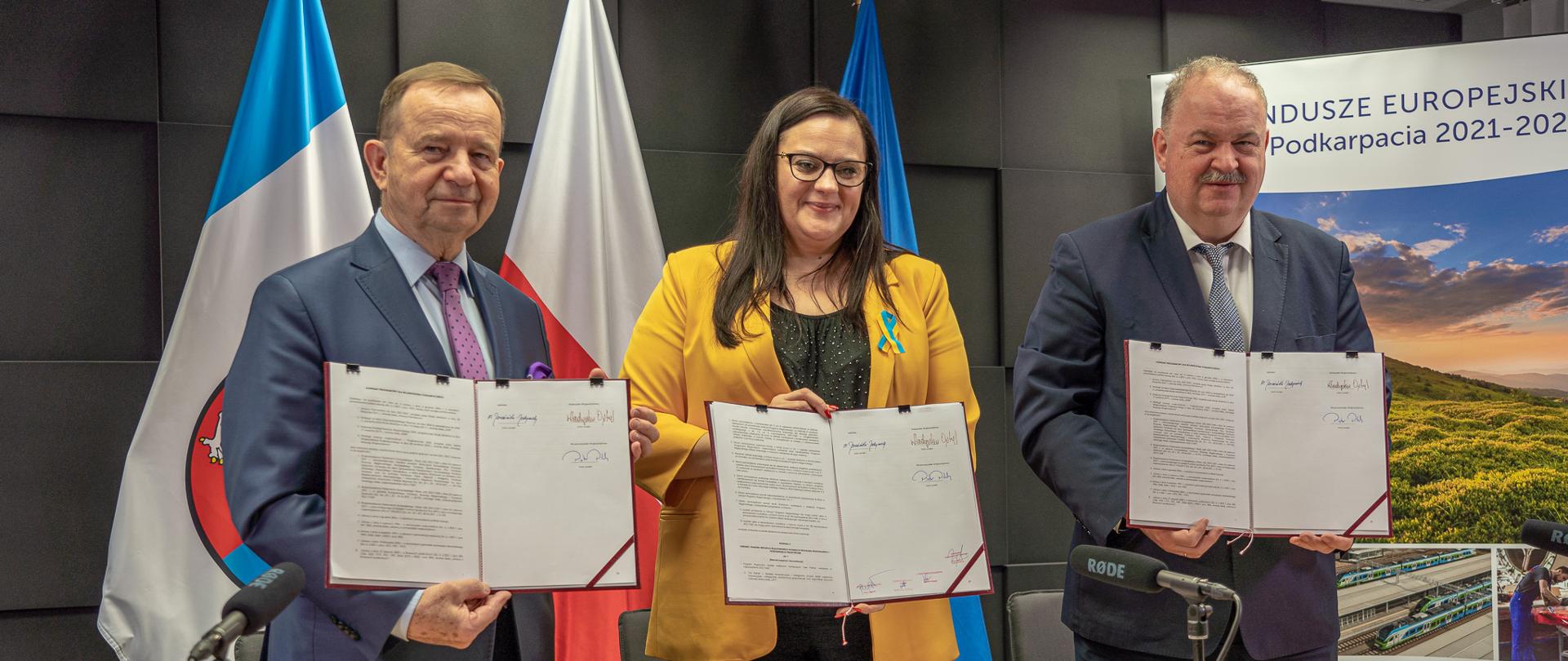 Trzy osoby pozują z dokumentami w rękach. Od lewej marszałek Władysław Ortyl, wiceminister Małgorzata Jarosińska-Jedynak oraz wicemarszałek Piotr Pilch. Za nimi flagi i ścianka.