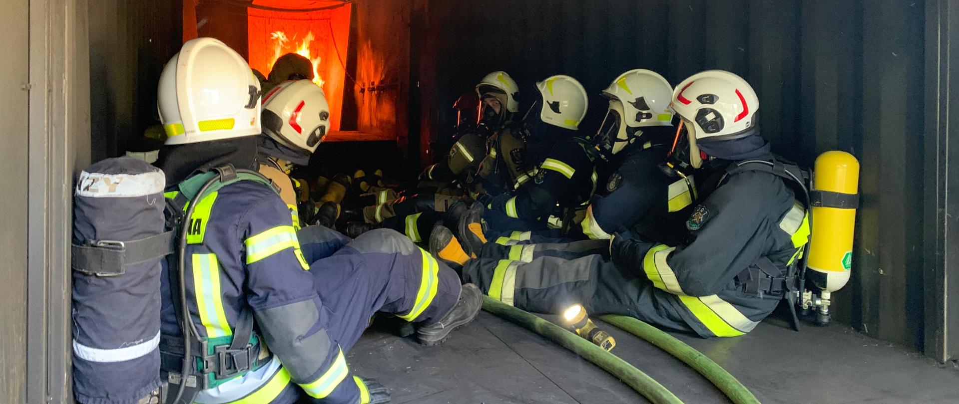 Strażacy OSP powiatu krapkowickiego na poligonie ratowniczym Komendy Wojewódzkiej PSP w Opolu uczestniczą w szkoleniu z gaszenia pożarów wewnętrznych. Strażacy siedzą w umundurowaniu bojowym i hełmach wewnątrz komory rozgorzenia. W tle widoczny ogień wewnątrz urządzenia ćwiczebnego.