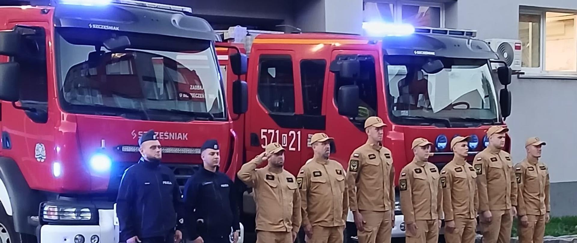 Strażacy oddają hołd dla zmarłych podczas akcji kolegów. Strażacy stoją na tle strażnicy.