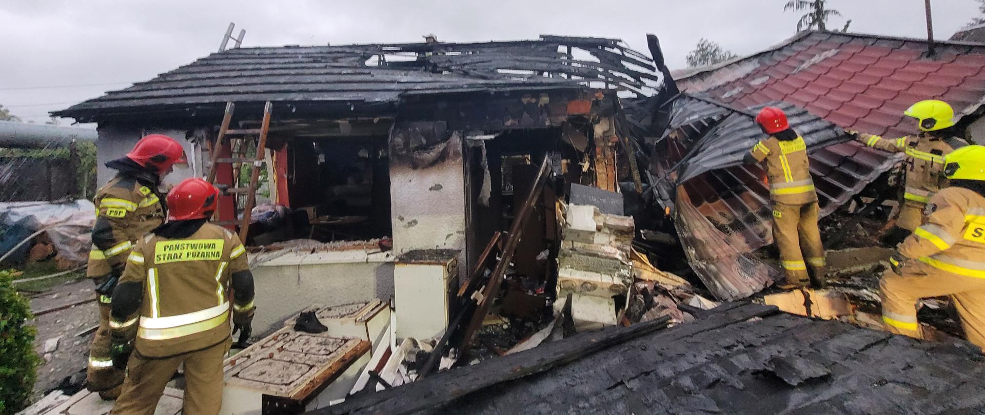 Uszkodzony budynek mieszkalny z czerwonym dachem. Przy zawalonej części pracuja strażacy w mundurach bojowych i hełmach.