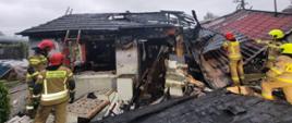 Uszkodzony budynek mieszkalny z czerwonym dachem. Przy zawalonej części pracuja strażacy w mundurach bojowych i hełmach.