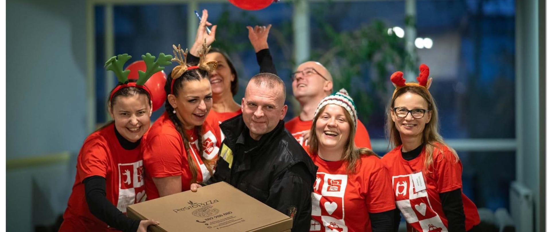 Przebrani wolontariusze w czerwonych koszulkach i strażak pozują rozbawieni do zdjęcia i trzymają kartony z pizzą