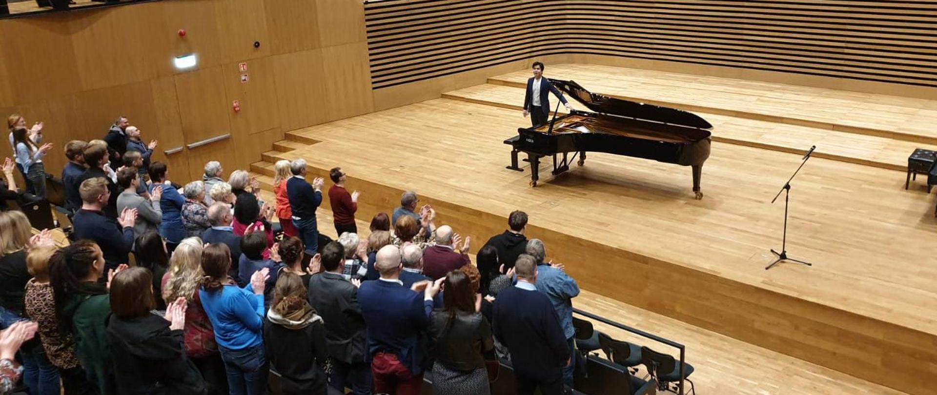 Pianista stoi przy fortepianie na scenie sali koncertowej PSM, z przodu publiczność klaszcze stojąc