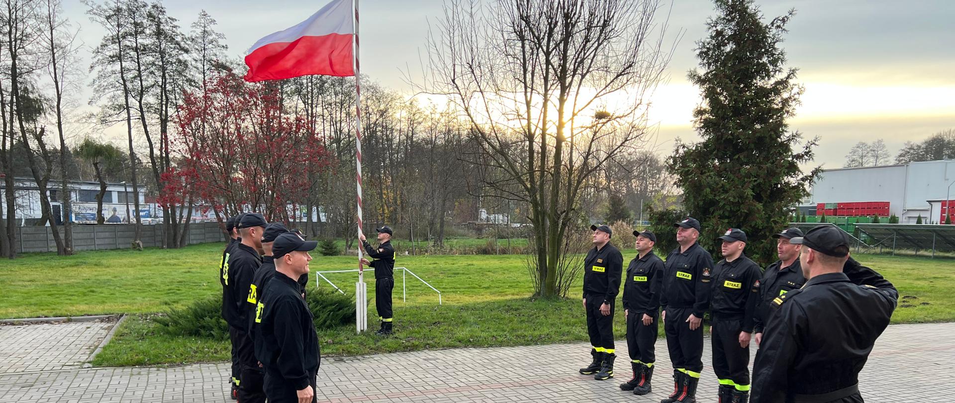 Zdjęcie przedstawia strażaków PSP podczas uroczystej zbiórki z okazji Narodowego Święta Niepodległości. Strażacy stoją w dwóch szeregach na przeciw siebie w umundurowaniu koszarowym. w oddali strażak wciągający flagę państwową na maszt