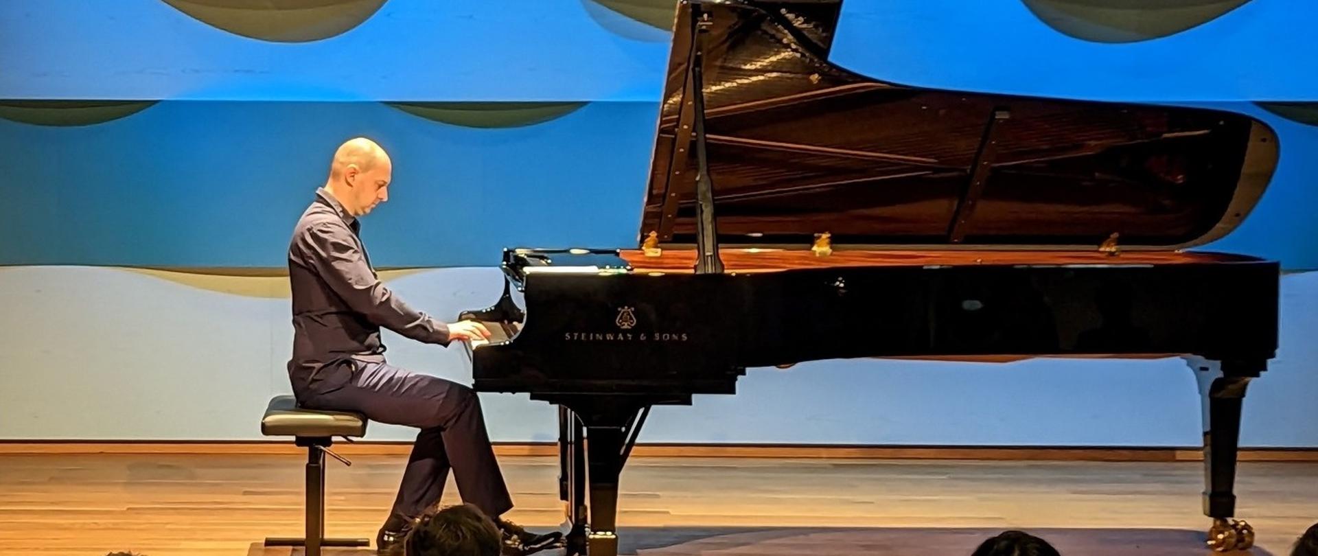 Zdjęcie mężczyzny grającego na fortepianie na scenie. W tle niebieska ściana o niejednolitej strukturze.