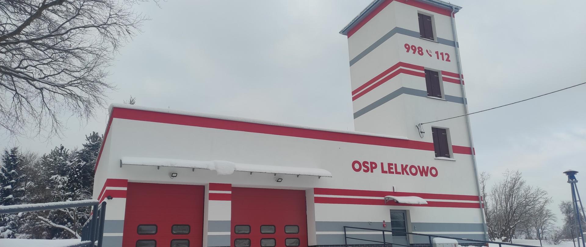 Na zdjęciu nowa remiza OSP Lelkowo, białe ściany z czerwonymi i szarymi pasami. Dwie duże czerwone bramy garażowe. Zimowa sceneria. 