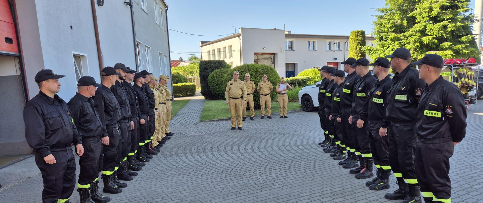 strażacy państwowej straży pożarnej podczas uroczystej zmiany służbowej stoją naprzeciw siebie w szeregu. pomiędzy nimi na środku stoi ubrany w mundur wyjściowy Komendant Powiatowy Państwowej Straży Pożarnej brygadier Marcin Mazurkiewicz.
