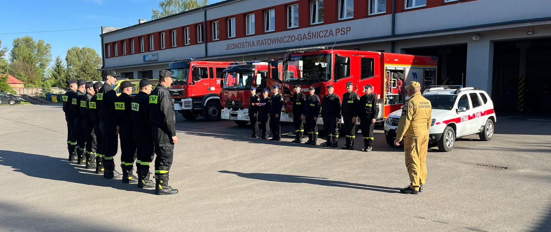 Na zdjęciu funkcjonariusze Państwowej Straży Pożarnej w umundurowaniu koszarowym ustawieni w dwóch szeregach twarzami do siebie między nimi funkcjonariusz w umundurowani służbowym. Obok jednego z szeregów ustawiony poczet flagowy z flagą Rzeczpospolitej Polskiej.