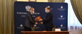 Zdjęcie przedstawia moment przekazania umowy. Za drewnianym stołem, na tle granatowej ścianki wizerunkowej Wojewody Mazowieckiego, stoi dwóch elegancko ubranych mężczyzn.