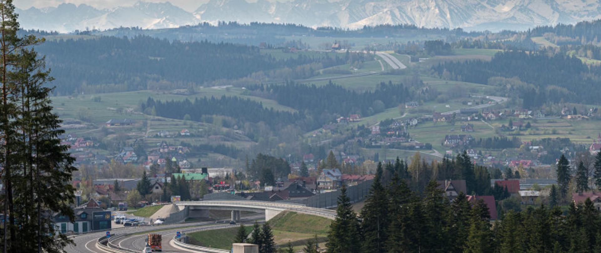 Droga krajowa z jadącymi po niej pojazdami. W tle zabudowania i malownicze góry.