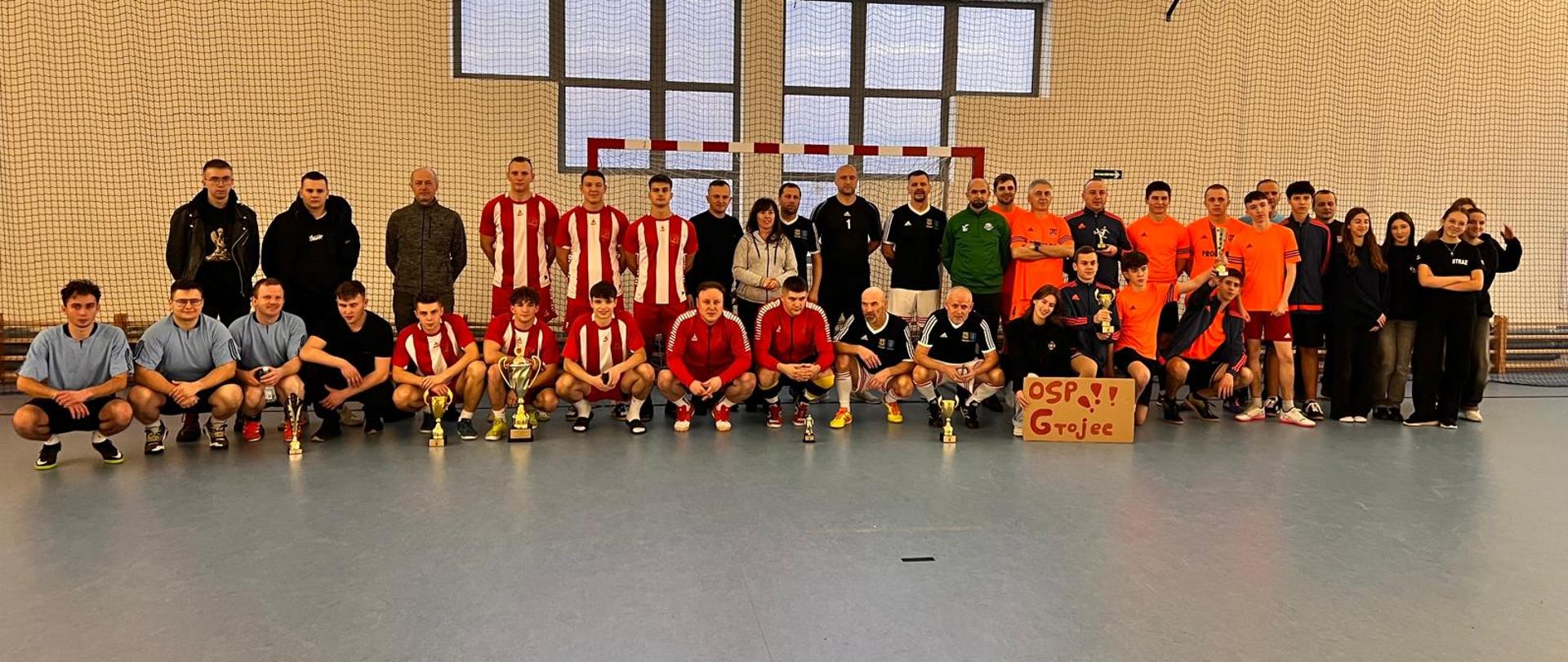 Na zdjęciu widać dużą grupę ludzi – uczestników XII Turnieju Służb Mundurowych w Alwerni. Stoją oni na hali sportowej, na tle bramki do piłki nożnej i siatki ochronnej. W centralnej części zdjęcia znajdują się drużyny sportowe w różnokolorowych strojach – w tym zawodnicy ubrani w czerwono-białe koszulki, czarne stroje oraz pomarańczowe komplety sportowe.
Na podłodze przed zawodnikami stoją cztery puchary, zdobyte podczas turnieju. Po prawej stronie zdjęcia grupa osób trzyma kartonowy plakat z napisem „OSP Grójec”. Wszyscy uczestnicy wyglądają na zadowolonych, uśmiechają się i pozują do wspólnego zdjęcia pamiątkowego.
W tle widać duże okna hali, przez które wpada naturalne światło, co nadaje zdjęciu jasną atmosferę.
