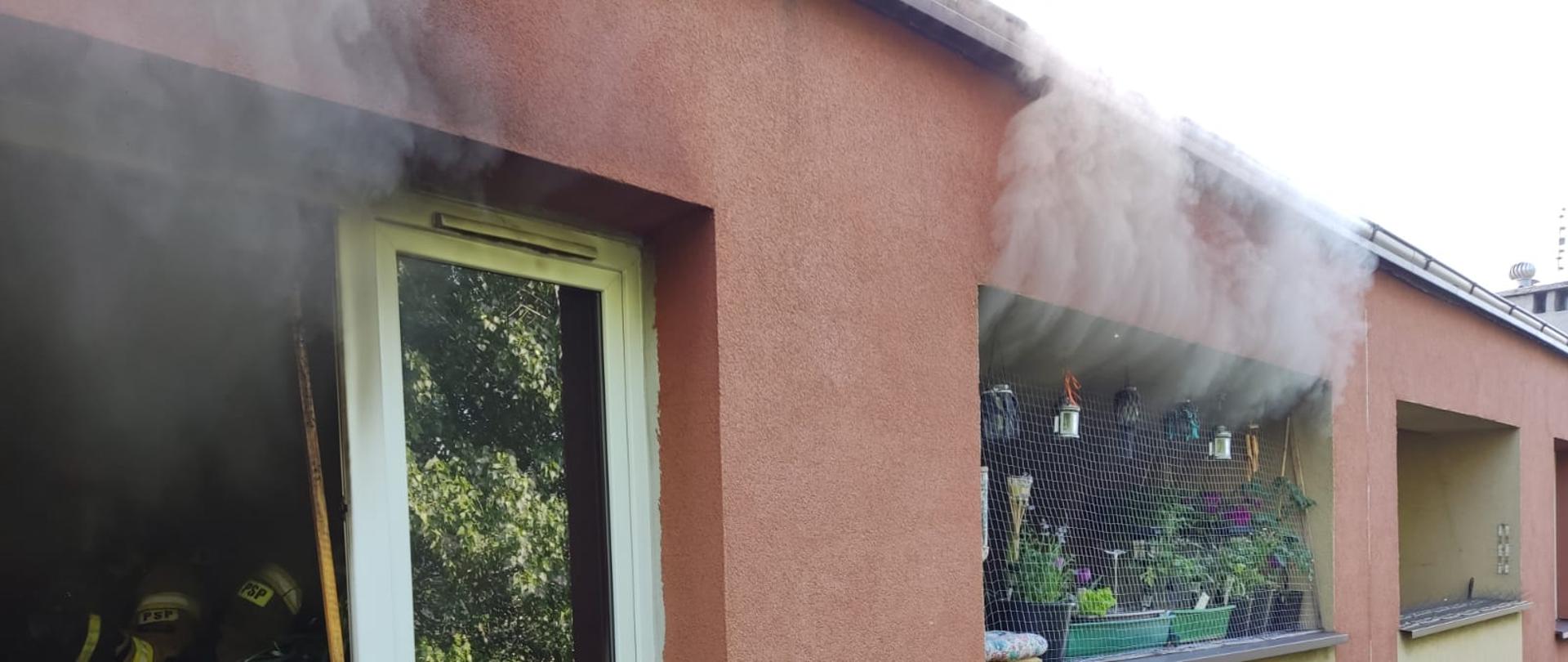 Zdjęcie przedstawia widok okna i balkonu na 4 piętrze z którego wydobywa się dym. Zdjęcie zrobione z zewnątrz budynku.