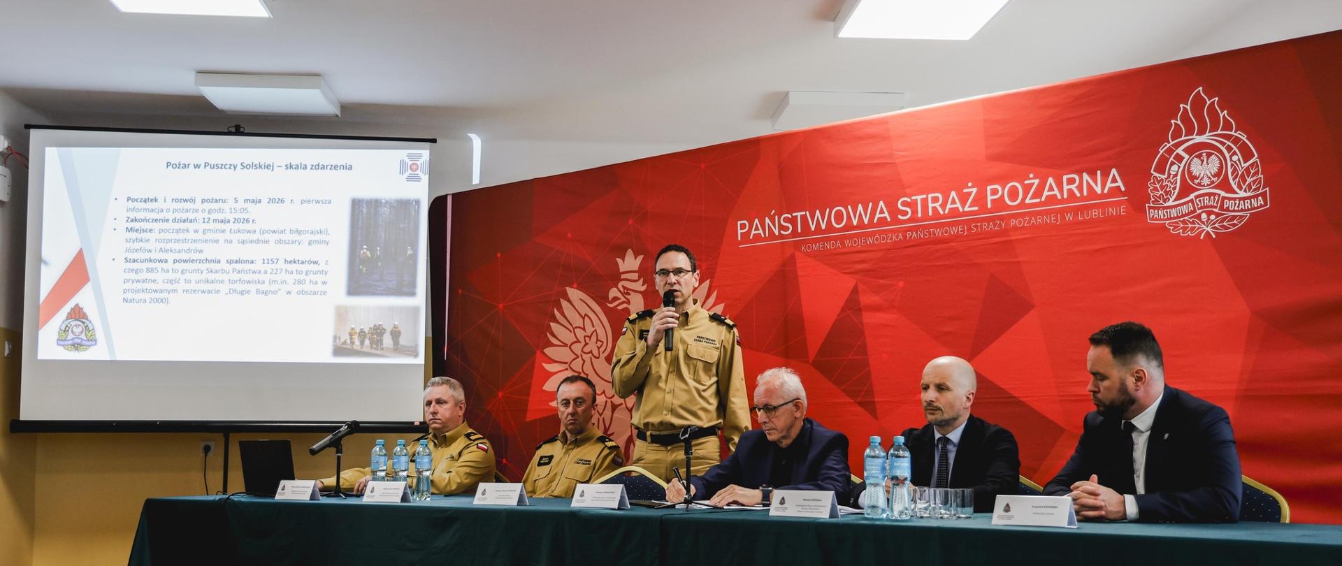 Przy stole prezydialnym podczas narady przemawia starszy mężczyzna w granatowej marynarce, trzymający mikrofon. Obok siedzą funkcjonariusze Państwowej Straży Pożarnej w umundurowaniu oraz przedstawiciele administracji. W tle widoczna czerwona ścianka z logotypem PSP.