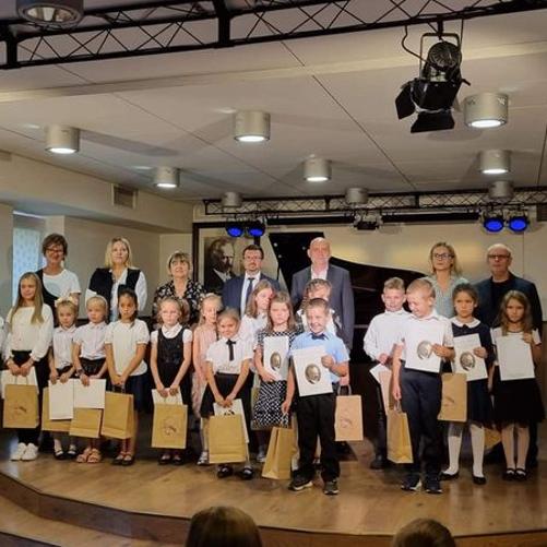 Fotografia przedstawia nowo przyjętych uczniów cyklu 6-letniego i nauczycieli. Wszyscy stoją na scenie auli szkoły. Z prawej strony widoczne 2 zasłonięte okna, za nauczycielami widać otwartą klapę od fortepianu, z tyłu na ścianie 4 niebieskie reflektory. Podłoga i boki w kolorze drewna. Na suficie 9 okrągłych lamp.