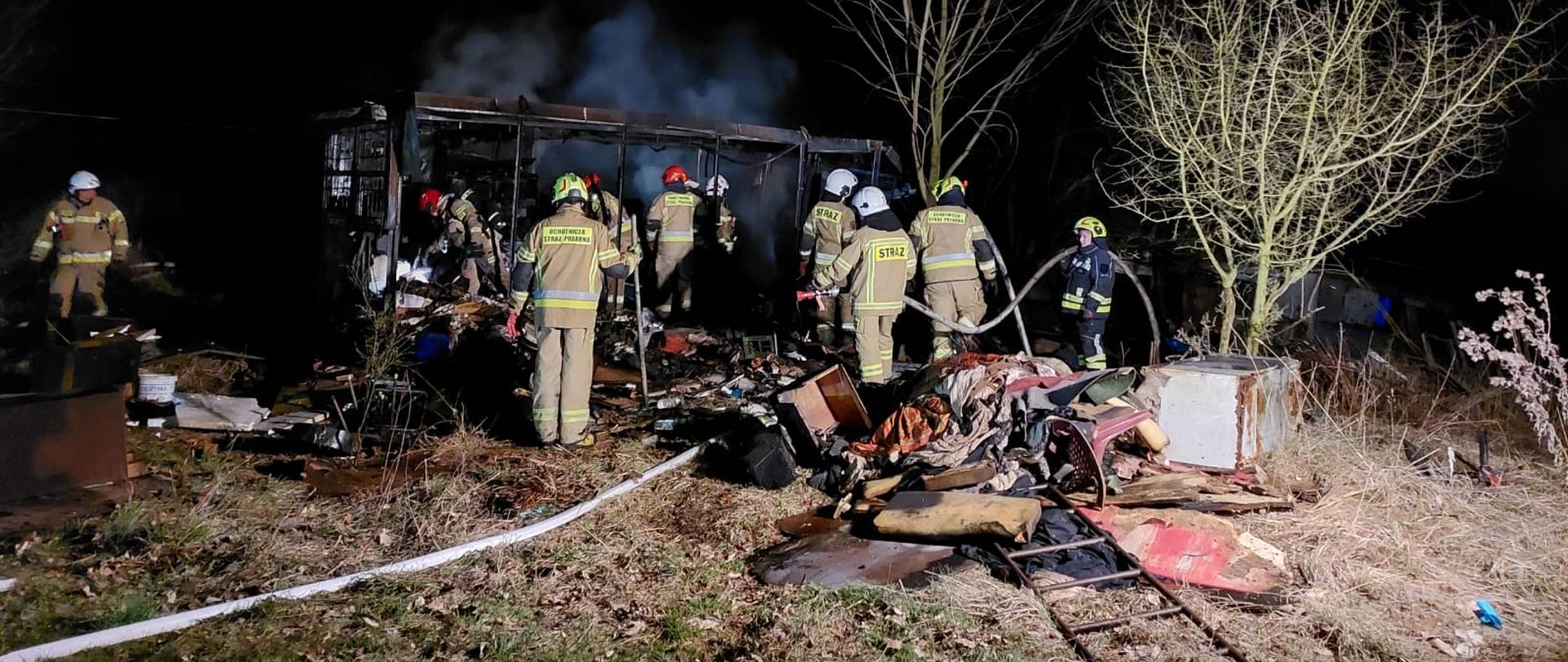 Zdjęcie przedstawia miejsce pożaru: ratownicy sprawdzają pogorzelisko, a z kontenera pozostał jedynie stalowy szkielet. 