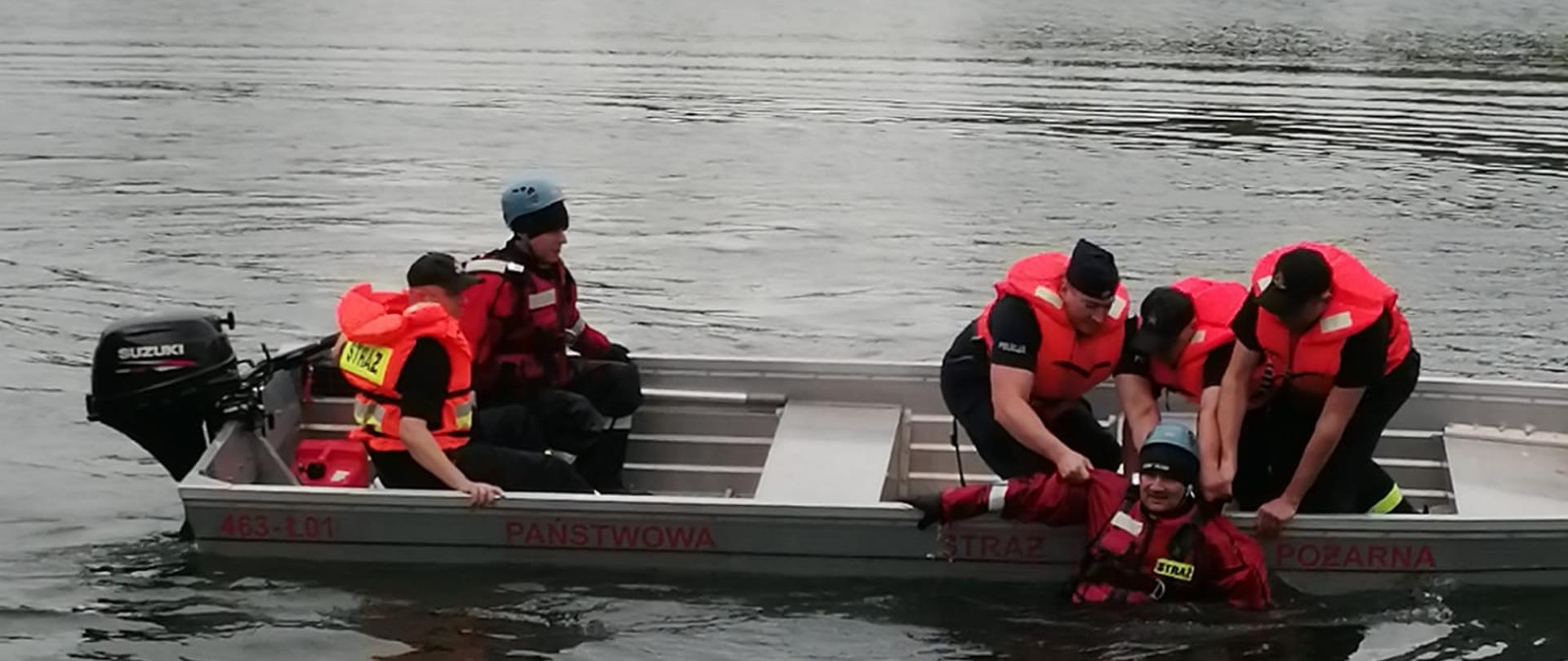 Widok na akwen wodny. W łodzi czterech strażaków oraz jeden funkcjonariusz policji ćwiczą ratowanie tonącego