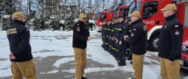 Pora zimowa, ośnieżony plac zewnętrzy komendy, zbiórka strażaków w dwuszeregu, w tle wystawione pojazdy pożarnicze. W oddali po prawej widoczny budynek Straży Pożarnej. 