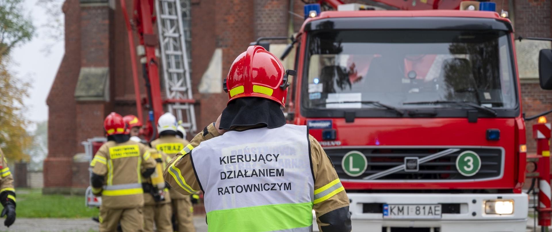 Na zdjęciu widoczny strażak kierujący ćwiczeniami z tyłu podnośnik SHP 25 oraz budynek kościoła