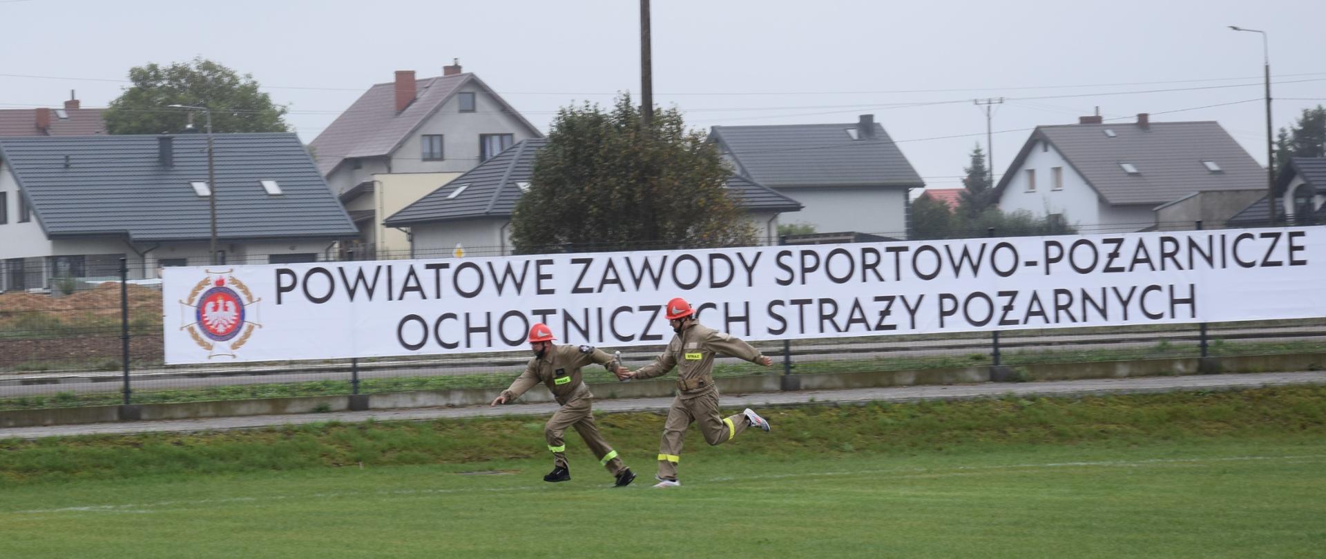 Dwóch strażaków OSP na boisku sportowym przekazuje sobie pałeczkę w biegu sztafetowym. Ubrani są w piaskowe mundury koszarowe oraz czerwone hełmy. Za nimi, na ogrodzeniu boiska widnieje duży baner z logo ZOSP RP napisem "POWIATOWE ZAWODY SPORTOWO-POŻARNICZE OCHOTNICZYCH STRAŻY POŻARNYCH". Za ogrodzeniem widać domy i pojedyncze drzewa, a nad niemi zachmurzone niebo. 