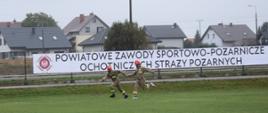 Dwóch strażaków OSP na boisku sportowym przekazuje sobie pałeczkę w biegu sztafetowym. Ubrani są w piaskowe mundury koszarowe oraz czerwone hełmy. Za nimi, na ogrodzeniu boiska widnieje duży baner z logo ZOSP RP napisem "POWIATOWE ZAWODY SPORTOWO-POŻARNICZE OCHOTNICZYCH STRAŻY POŻARNYCH". Za ogrodzeniem widać domy i pojedyncze drzewa, a nad niemi zachmurzone niebo. 