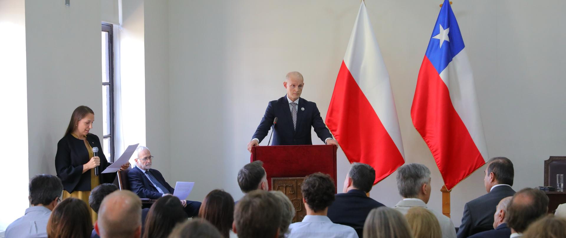 Na zdjęciu jest mężczyzna w garniturze, który stoi przy mównicy. Za nim są dwie flagi: Polski i Chile. Po lewej stronie jest kobieta z mikrofonem. Przed nimi jest grupa osób siedzących na krzesłach. Są tyłem do obiektywu. 