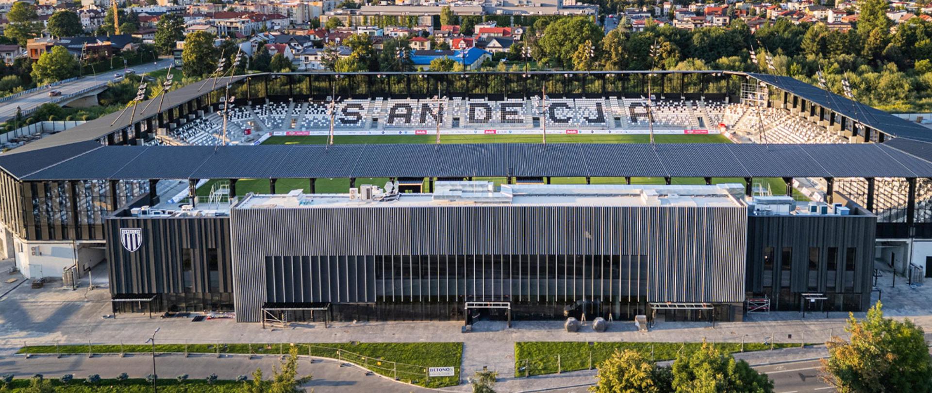 Na zdjęciu z lotu ptaka widać nowo wybudowany stadion. Widoczna jest murawa oraz trybuny. Na trybunach widoczny napis „Sandecja” – nazwa lokalnego klubu piłkarskiego. 