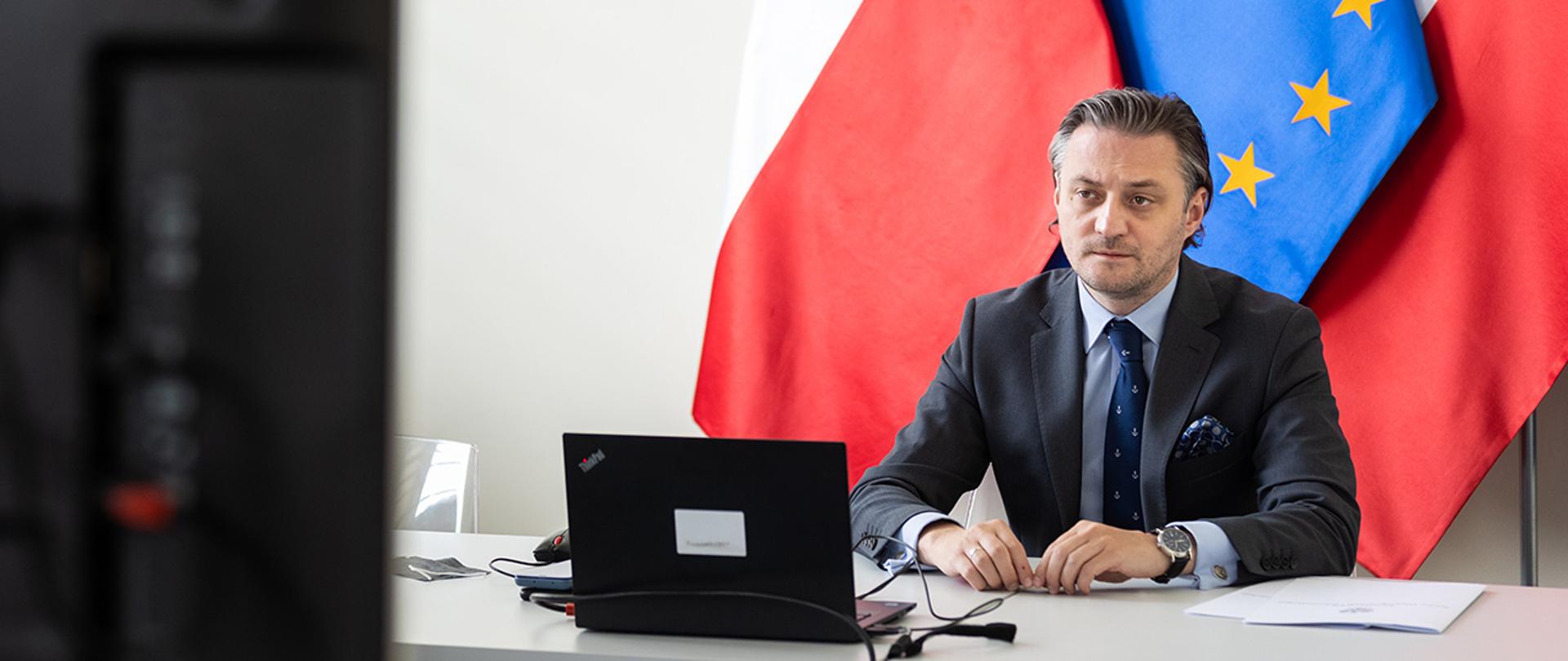 Na zdjęciu widać wiceministra Bartosza Grodeckiego siedzącego za stołem, przed ekranem na tle flag Polski i UE.