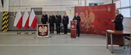 Na garażu obok flag Polski i banera Państwowej Straży Pożarnej stoją w rzędzie strażacy ubrani w mundury galowe. Przed nimi wystawione jest Godło Polski
